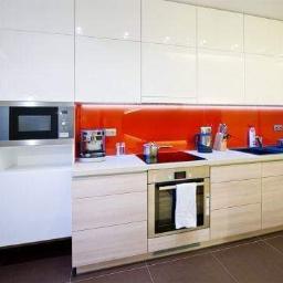 Porta Lis - Nowoczesna kuchnia z białymi, błyszczącymi szafkami górnymi i jasnymi, drewnopodobnymi szafkami dolnymi, z pomarańczowym panelem szklanym nad blatem, wyposażona w wbudowany piekarnik z ręcznikiem...