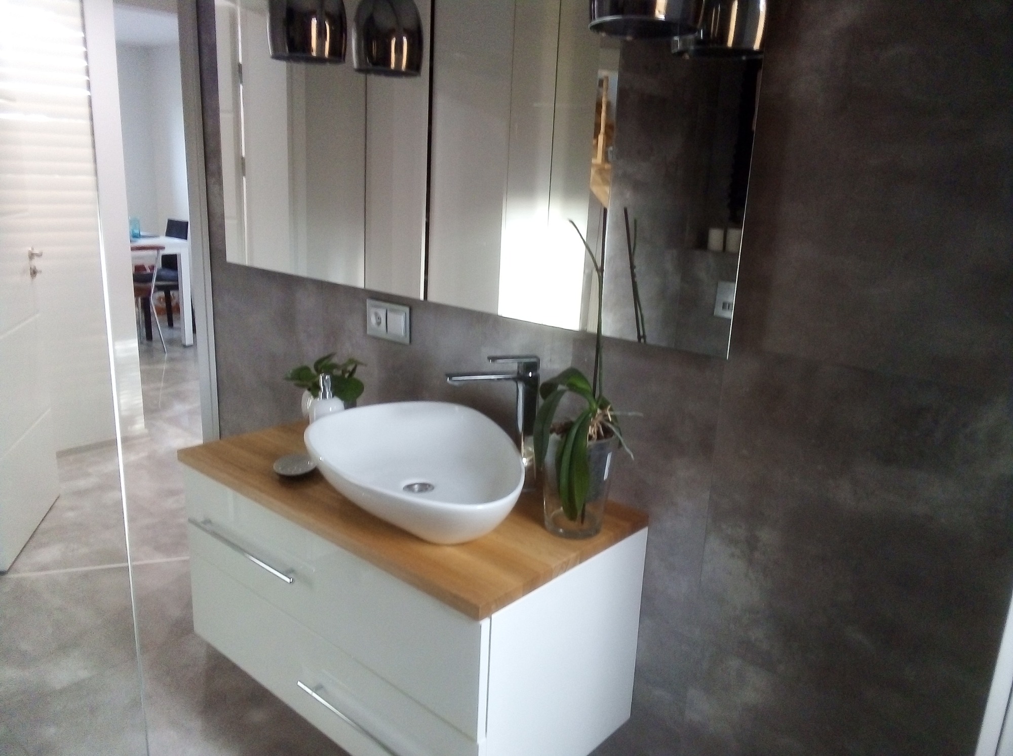 Nowoczesna łazienka z białą, owalną umywalką nablatową na szafce z drewnianym blatem, szara ściana i duże lustro z oświetleniem.