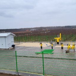 MAGBUD Magdalena Ostrowska - Ogrodzony teren z żółtą instalacją gazową i budynkiem technicznym w Skierniewicach. Kostka brukowa na podłożu, w tle sad. Widok z góry.