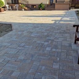 MAGBUD Magdalena Ostrowska - Ułożona kostka brukowa w odcieniach szarości i beżu tworzy wzorzysty chodnik przed domem. Widoczne schody wejściowe i elementy architektury ogrodowej.