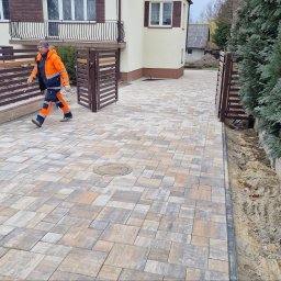 MAGBUD Magdalena Ostrowska - Ułożona kostka brukowa na podjeździe z otwartą bramą; widoczny pracownik w odblaskowym ubraniu, budynek z balkonem i fragment ogrodu z iglakami.