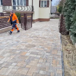 Ułożona kostka brukowa na podjeździe z otwartą bramą; widoczny pracownik w odblaskowym ubraniu, budynek z balkonem i fragment ogrodu z iglakami.