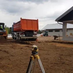 Prace ziemne na placu budowy: koparko-ładowarka przy pryzmie ziemi, pomarańczowa wywrotka w tle, niwelator na statywie na pierwszym planie, w tle budynek w stanie surowym otwartym.