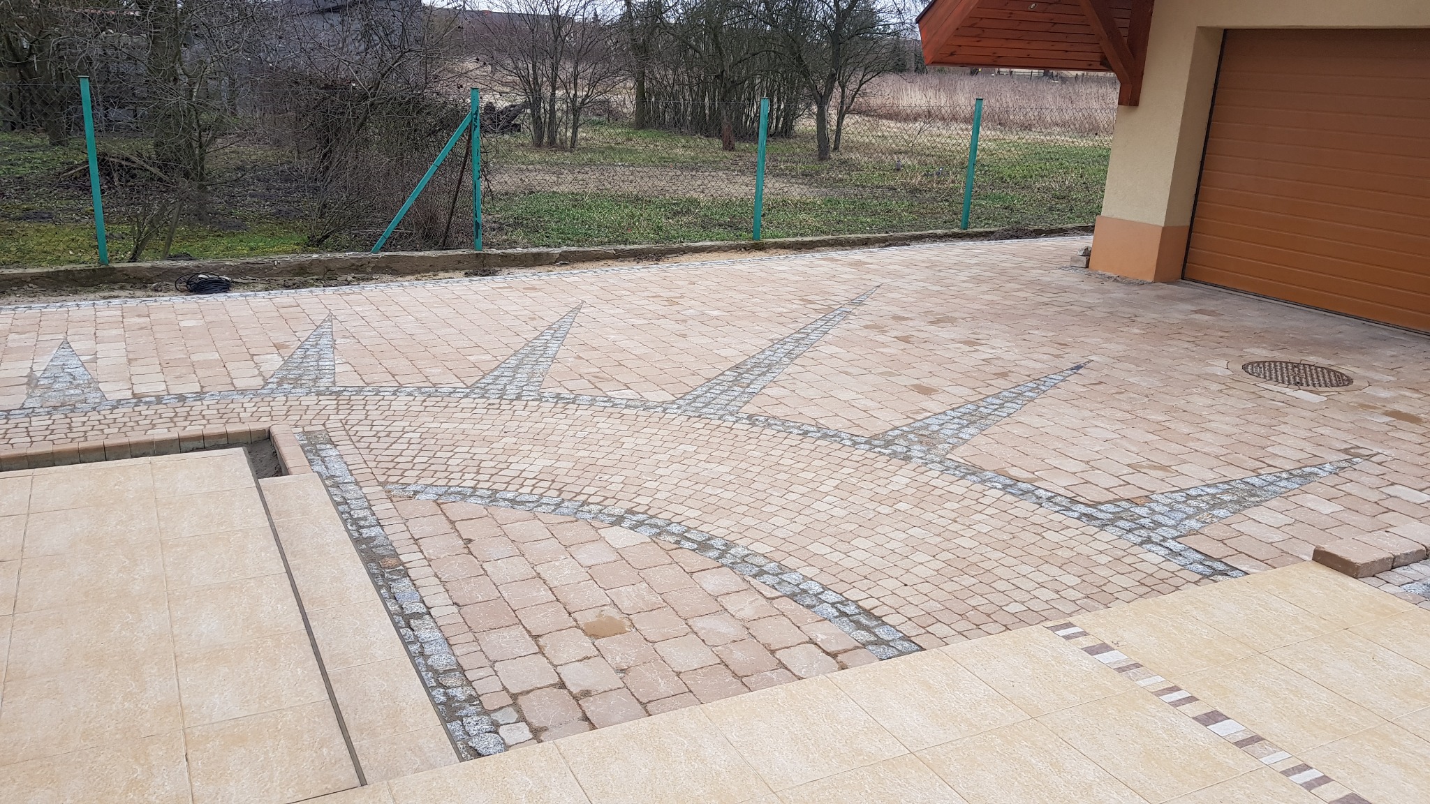 Beżowa kostka brukowa ułożona w oryginalny wzór rozety w pobliżu brązowej bramy garażowej, z widocznymi schodami wejściowymi wyłożonymi płytkami.