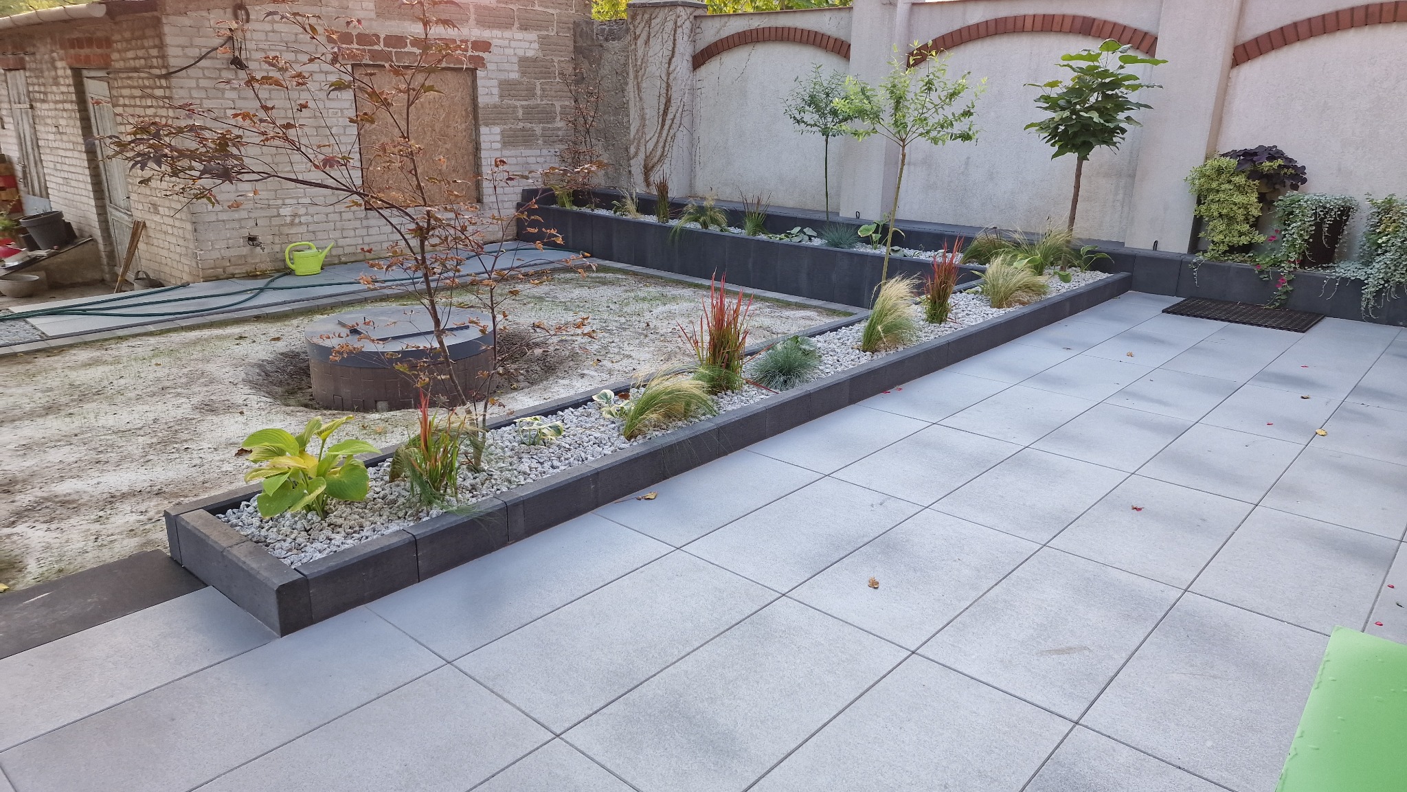 Nowoczesny taras z szarej kostki, obrzeża z kostki granitowej, rabaty z roślinami ozdobnymi i drzewkami. W tle mur z dekoracyjnym łukiem z cegły.