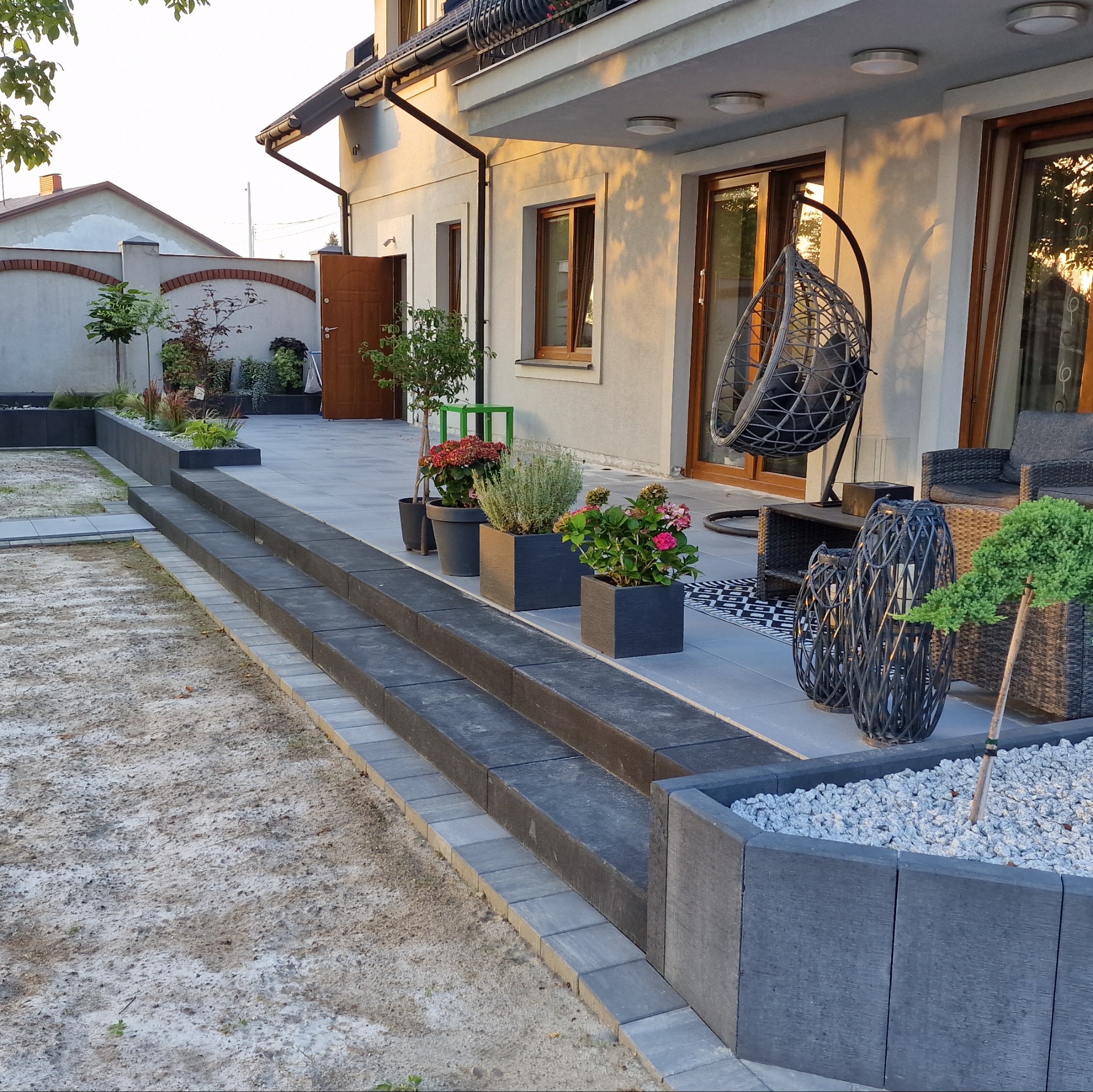 Nowoczesny taras z kostki, schody z donicami, wiszące krzesło. Starannie ułożona nawierzchnia, rośliny w donicach. Elementy dekoracyjne i meble ogrodowe.