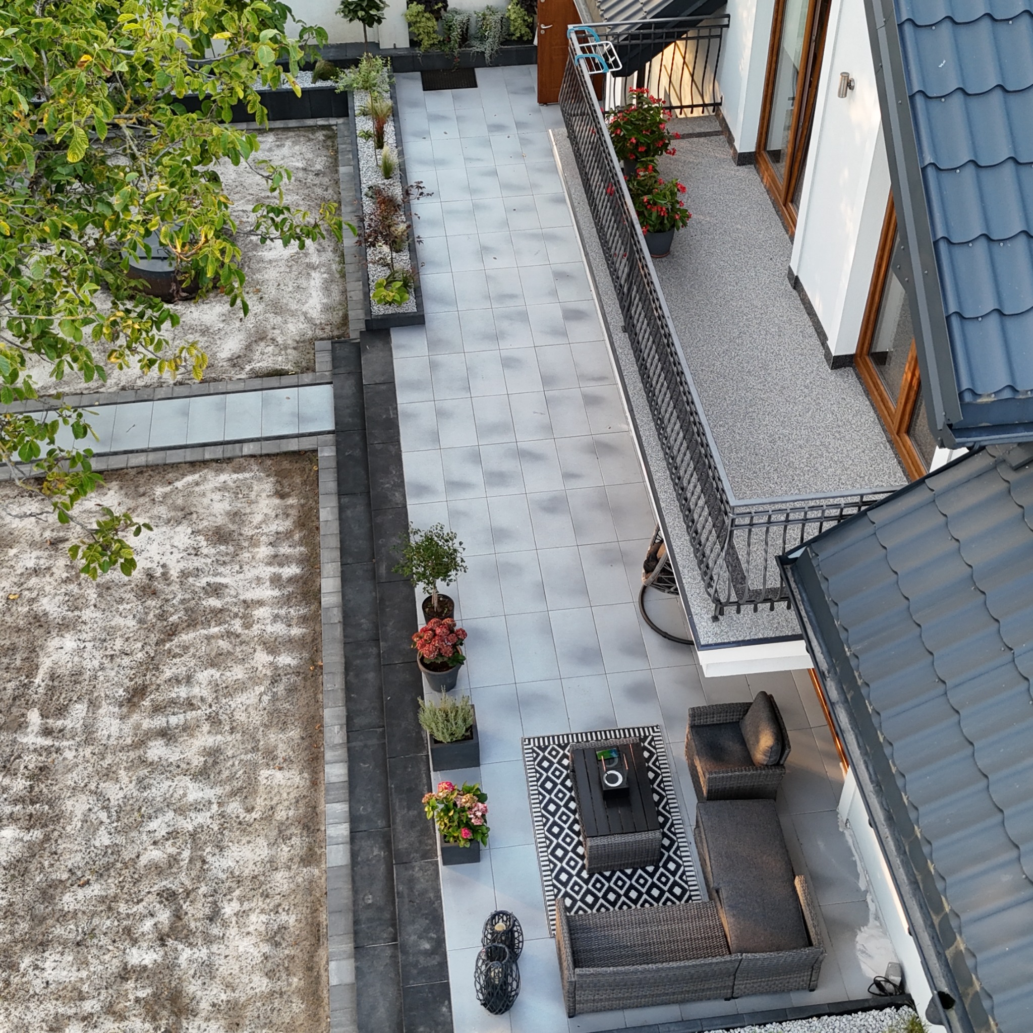 Z góry widok na ułożoną, jasnoszarą kostkę brukową tworzącą taras i ścieżkę w ogrodzie, z meblami ogrodowymi i roślinami w donicach.