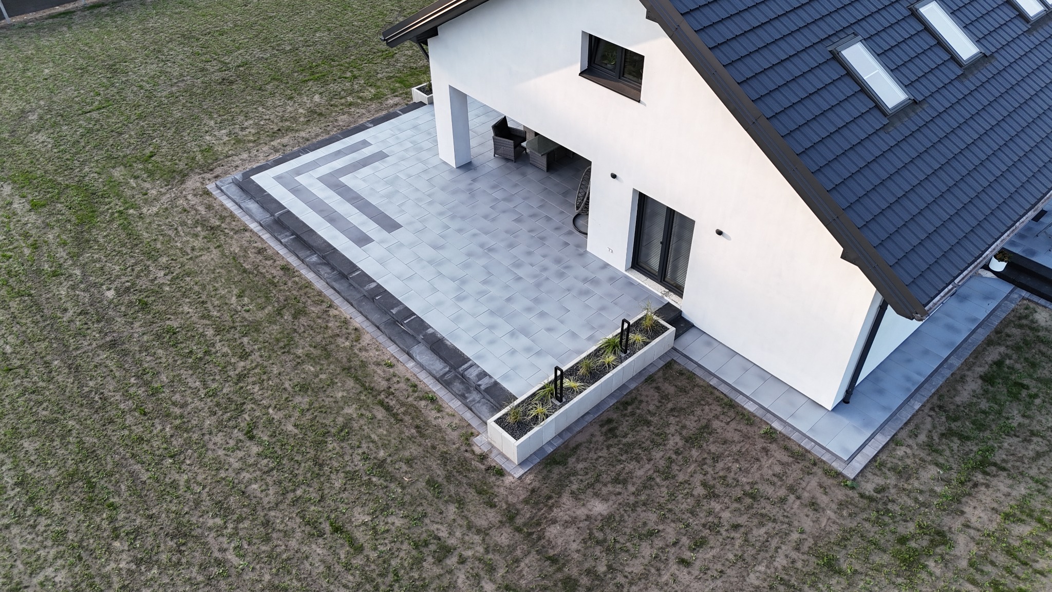 Nowoczesny taras z szarej kostki brukowej ułożonej we wzór, widok z góry, z przyległym trawnikiem i fragmentem białego domu z ciemną dachówką.