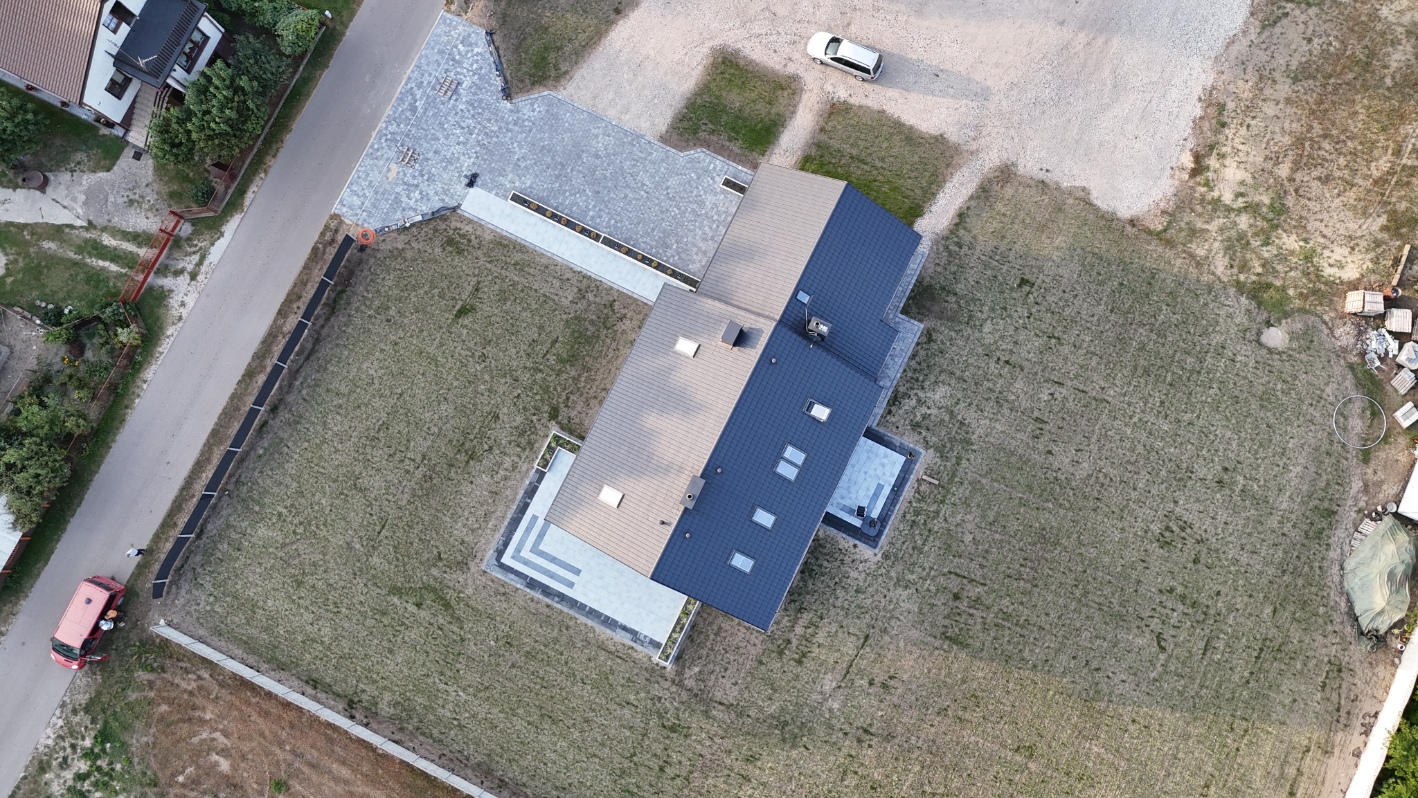 Widok z drona na dom z ciemnym dachem, jasną kostką brukową przed wejściem i podjazdem. Otoczenie to trawnik i droga dojazdowa z zaparkowanym autem.
