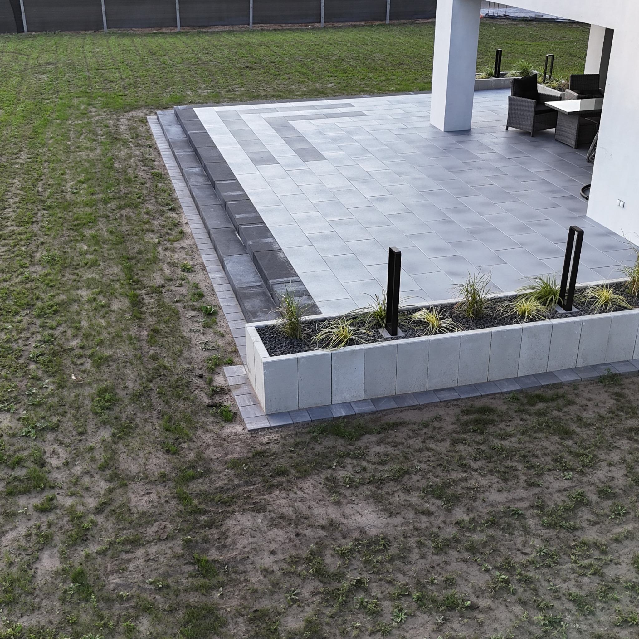 Nowoczesny taras z szarej kostki brukowej, z donicą z roślinami i oświetleniem, widok z góry. Umeblowany fragment tarasu z białym słupem w tle.