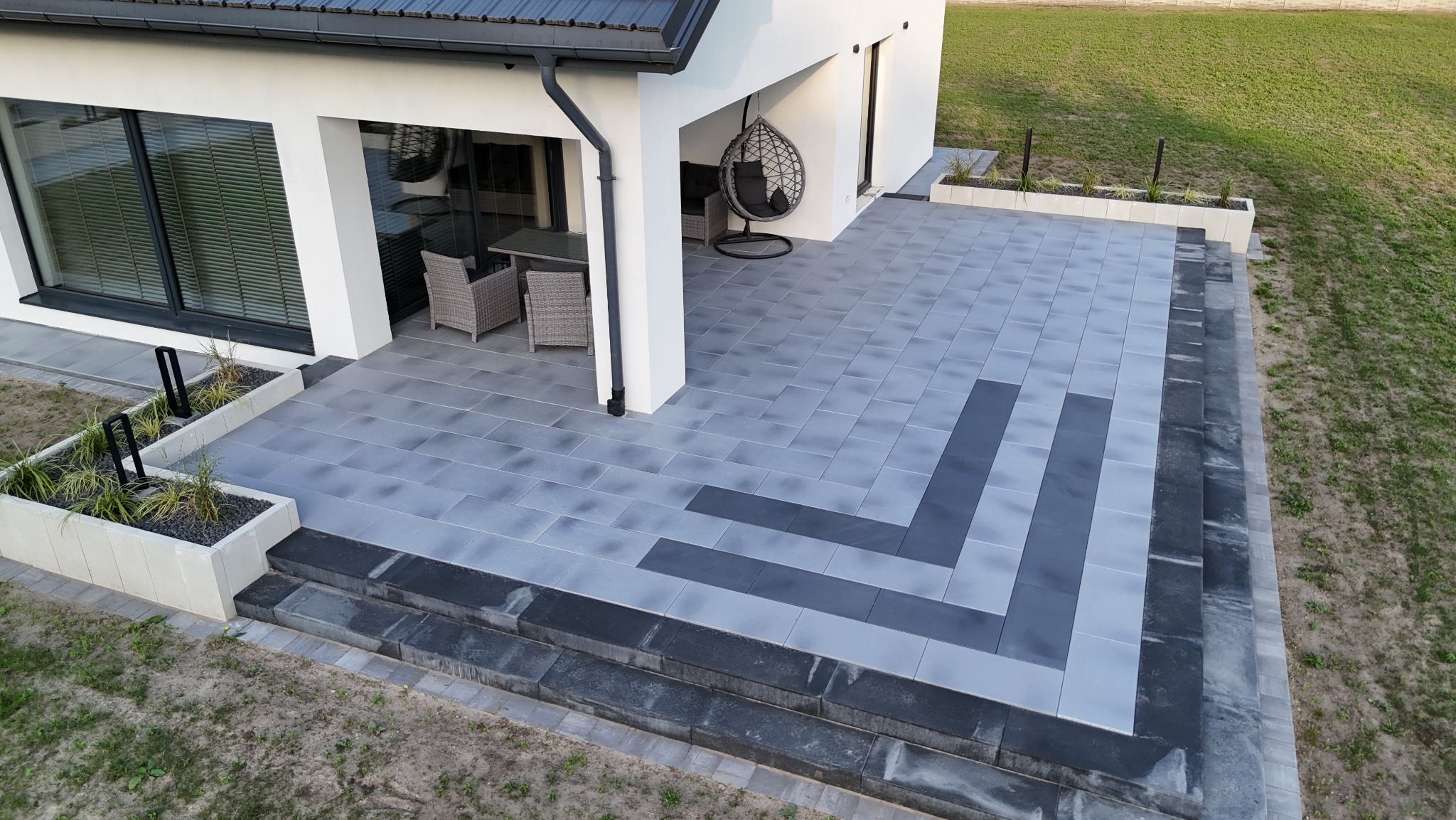 Nowoczesny taras z szarej kostki brukowej ułożonej we wzór z ciemniejszymi pasami. Widoczne schody, meble ogrodowe i elementy dekoracyjne.