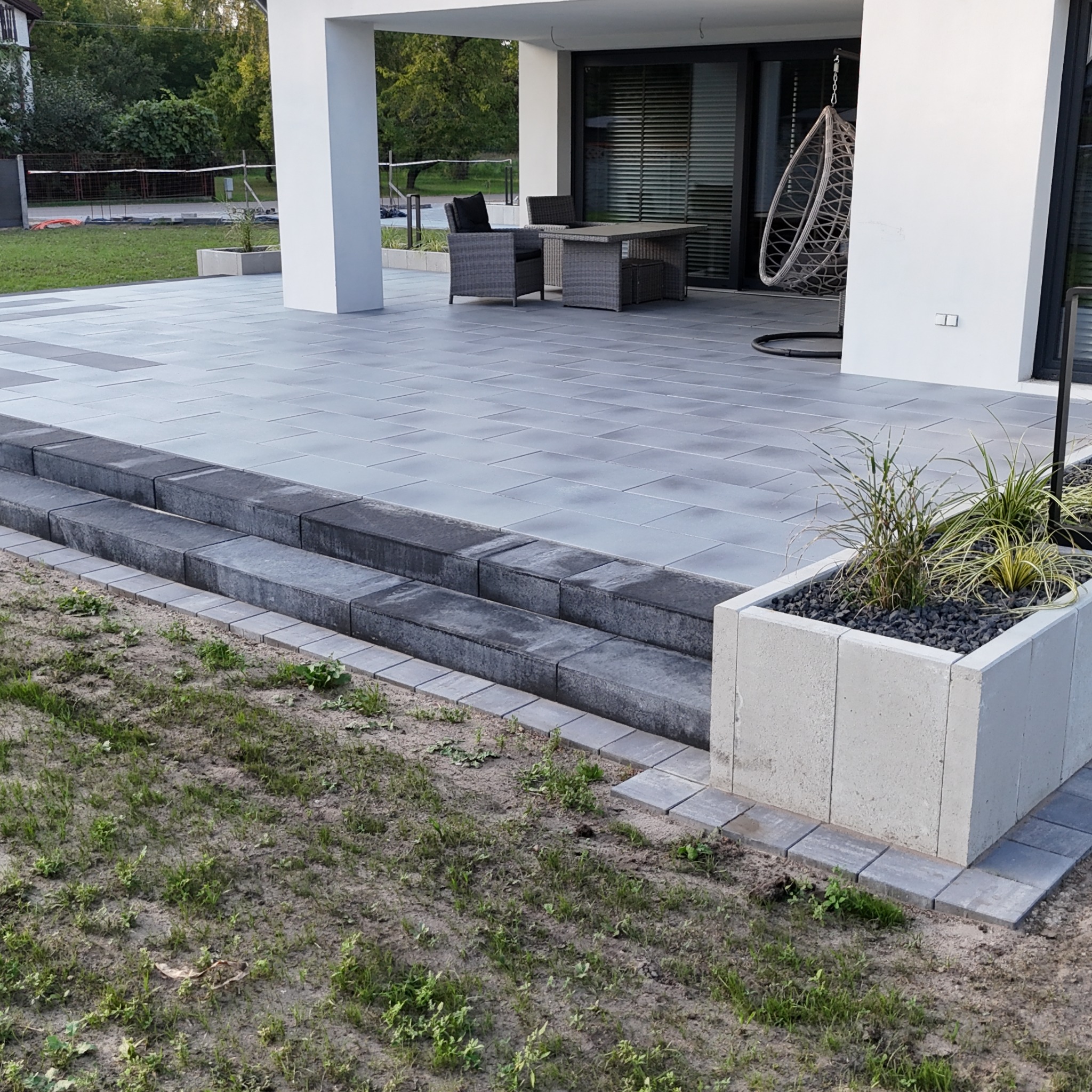 Nowoczesny taras z szarych płyt, schody z kostki granitowej i betonowa donica z roślinami. Widok na dom z meblami ogrodowymi i wiszącym fotelem.