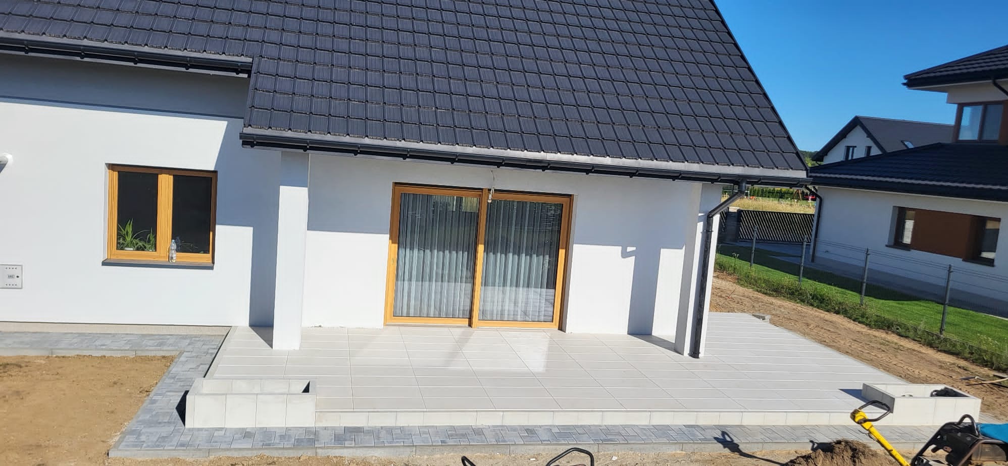 Jasna, nowa elewacja domu z tarasem wyłożonym płytkami i kostką. Widoczne detale wykończenia, okno z rośliną i sąsiednie budynki w słoneczny dzień.