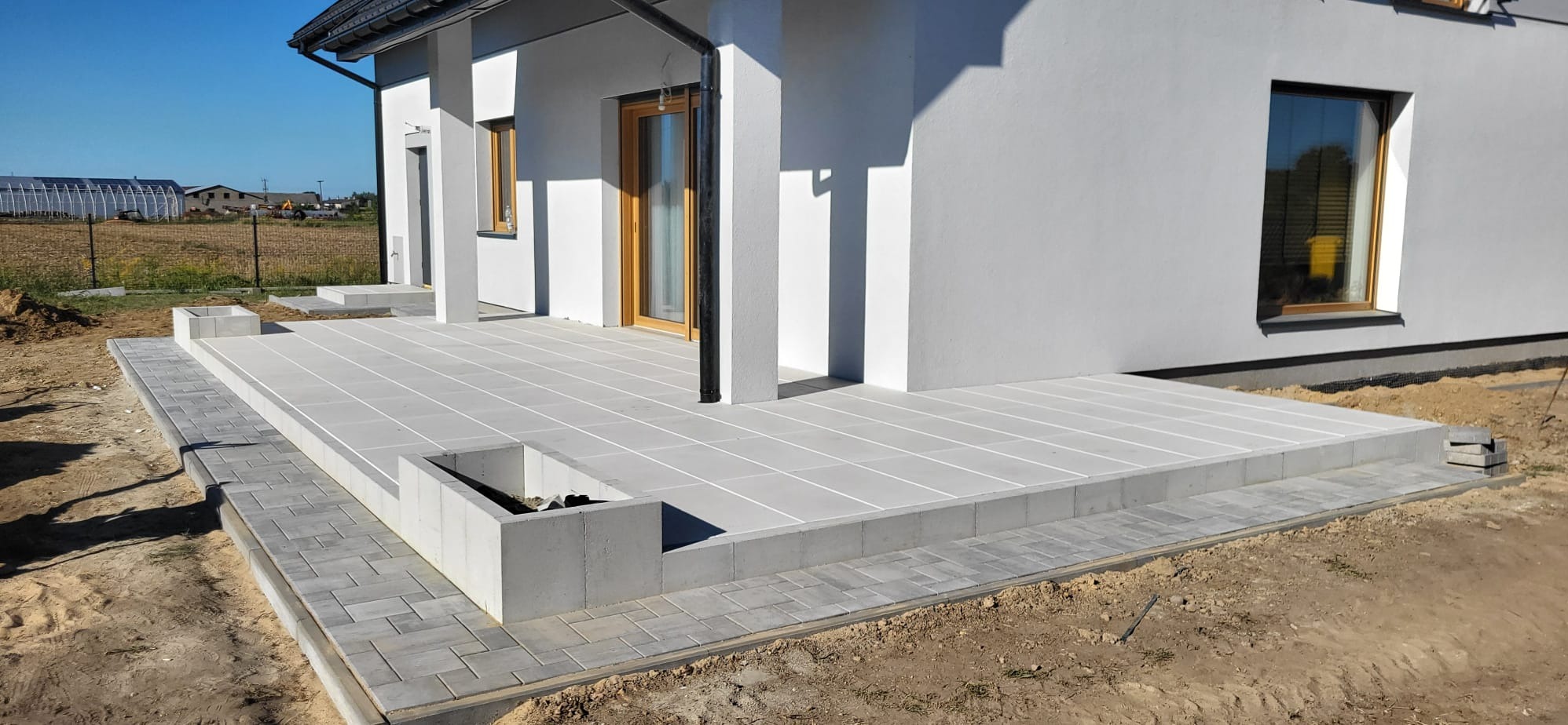 Nowoczesny taras z jasnoszarej kostki brukowej o geometrycznym wzorze, przylegający do białego budynku z drewnianymi oknami. W tle widoczne pole i zabudowania.