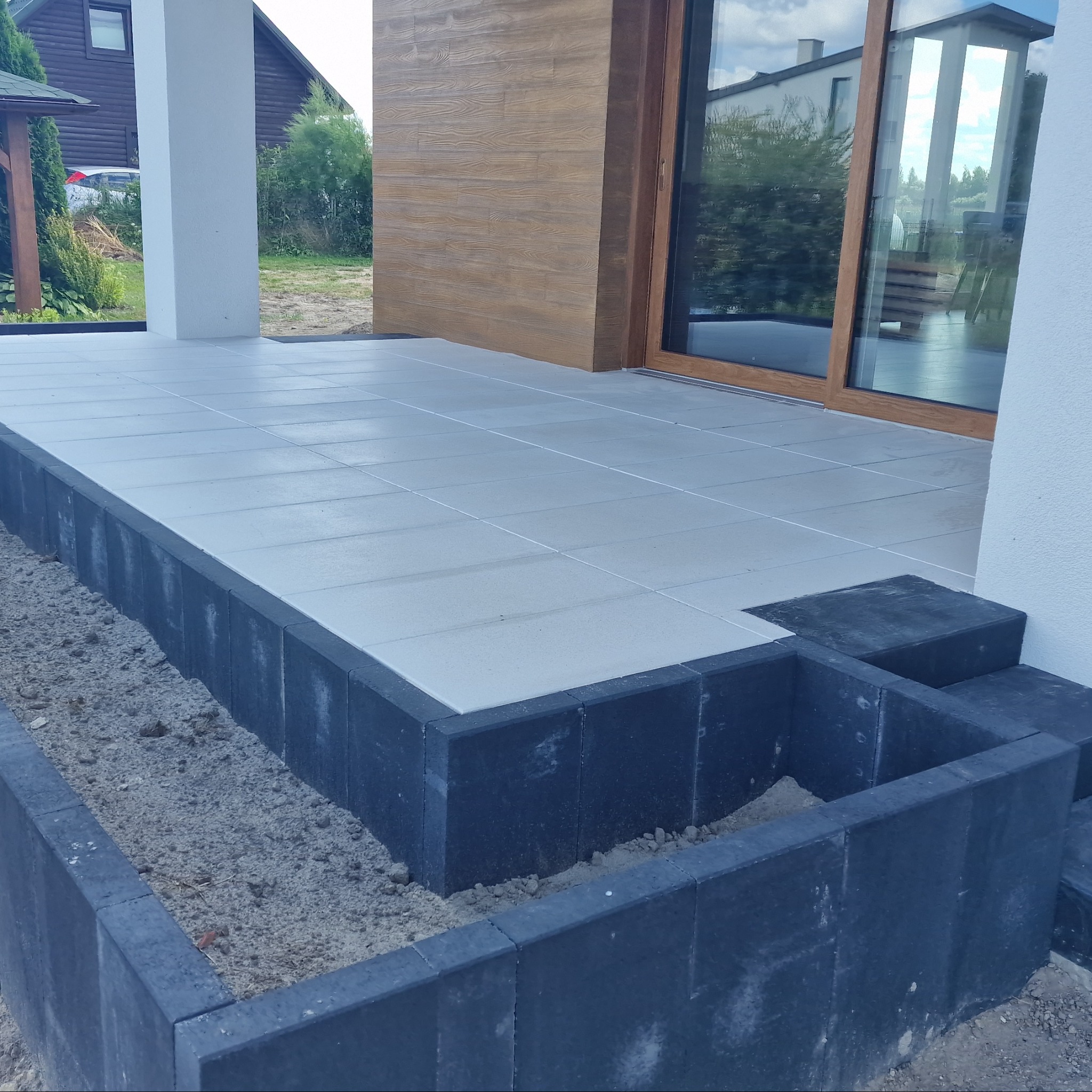 Jasnoszara kostka brukowa na tarasie z ciemnymi obrzeżami i schodami, widok z dołu, w tle okno tarasowe i fragment domu.