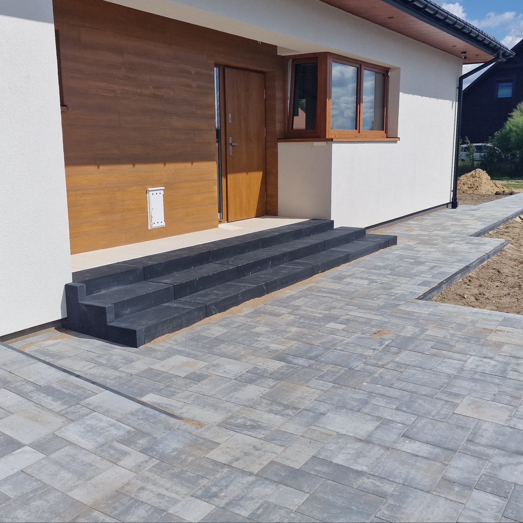 Ułożona szara kostka brukowa prowadząca do drzwi wejściowych z trzema czarnymi stopniami. Widoczne wykończenie domu w nowoczesnym stylu.