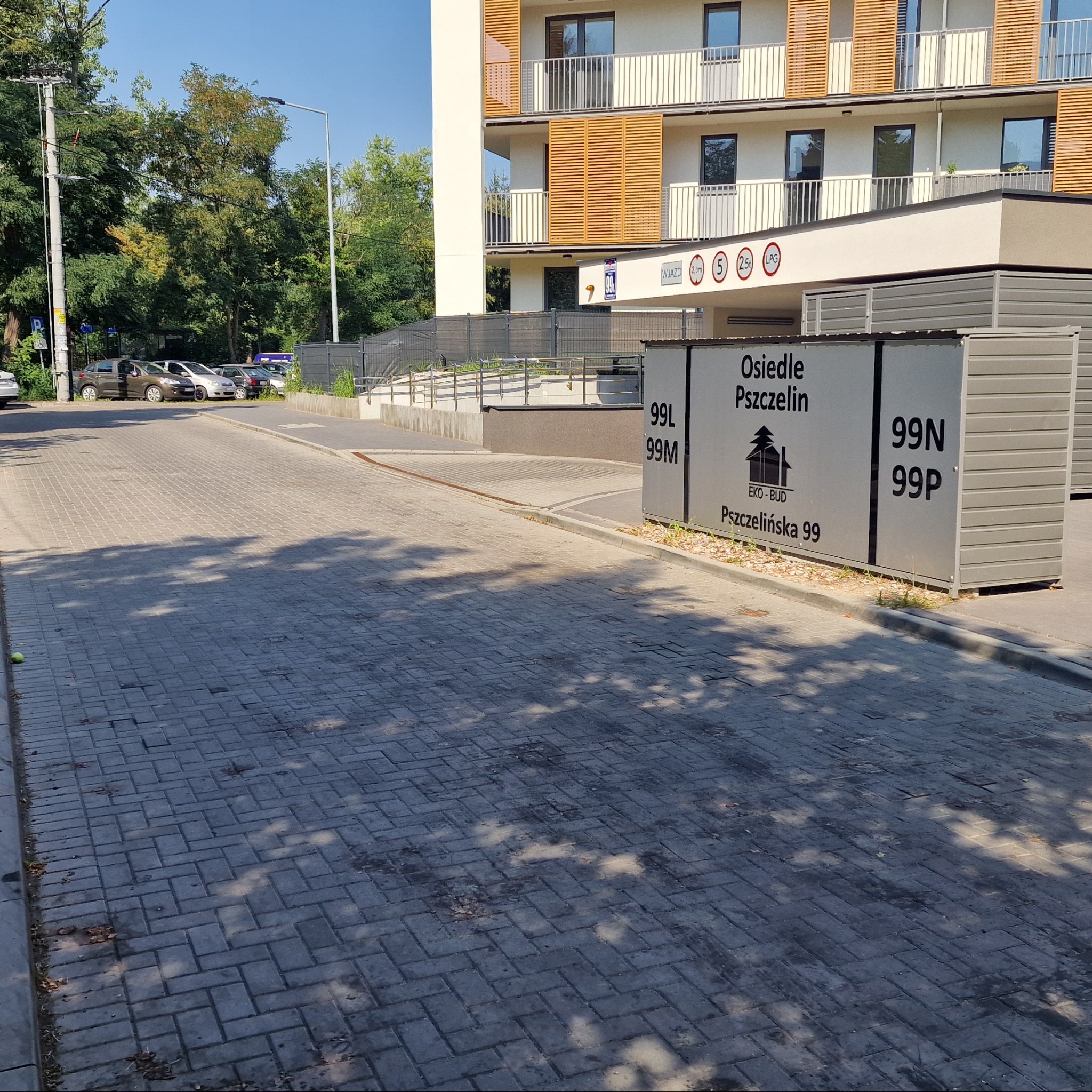 Szara kostka brukowa na podjeździe osiedla Pszczelin, z widocznymi miejscami parkingowymi i budynkiem w tle. Cień drzew na nawierzchni.