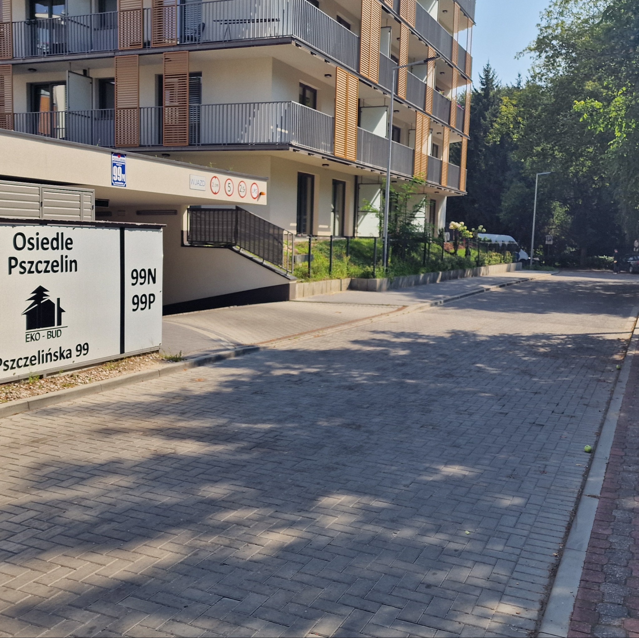 Nowoczesny budynek mieszkalny z balkonami, widok z poziomu ulicy wyłożonej szarą kostką brukową. Tablica informacyjna z nazwą osiedla po lewej.