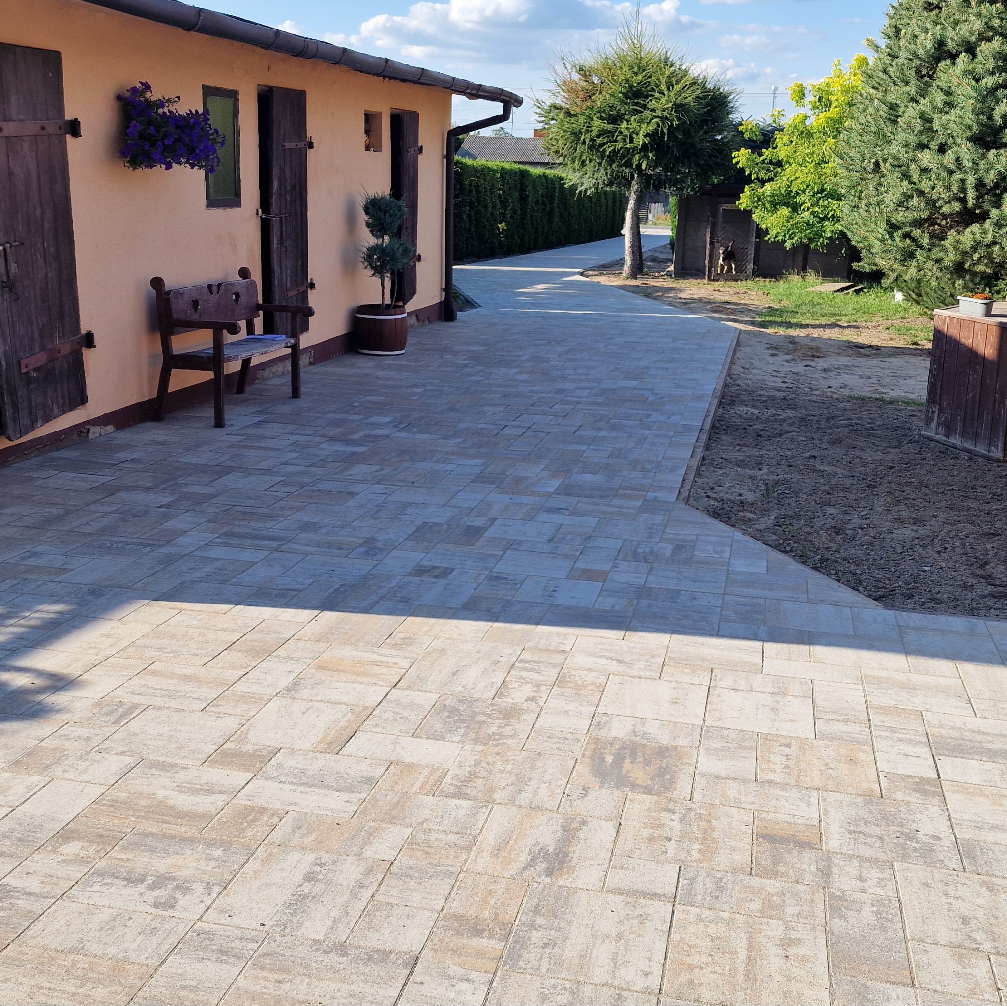 Jasna kostka brukowa ułożona wzdłuż budynku z drewnianymi okiennicami i ławką. Widoczne krzewy i drzewa w tle. Słoneczny dzień.