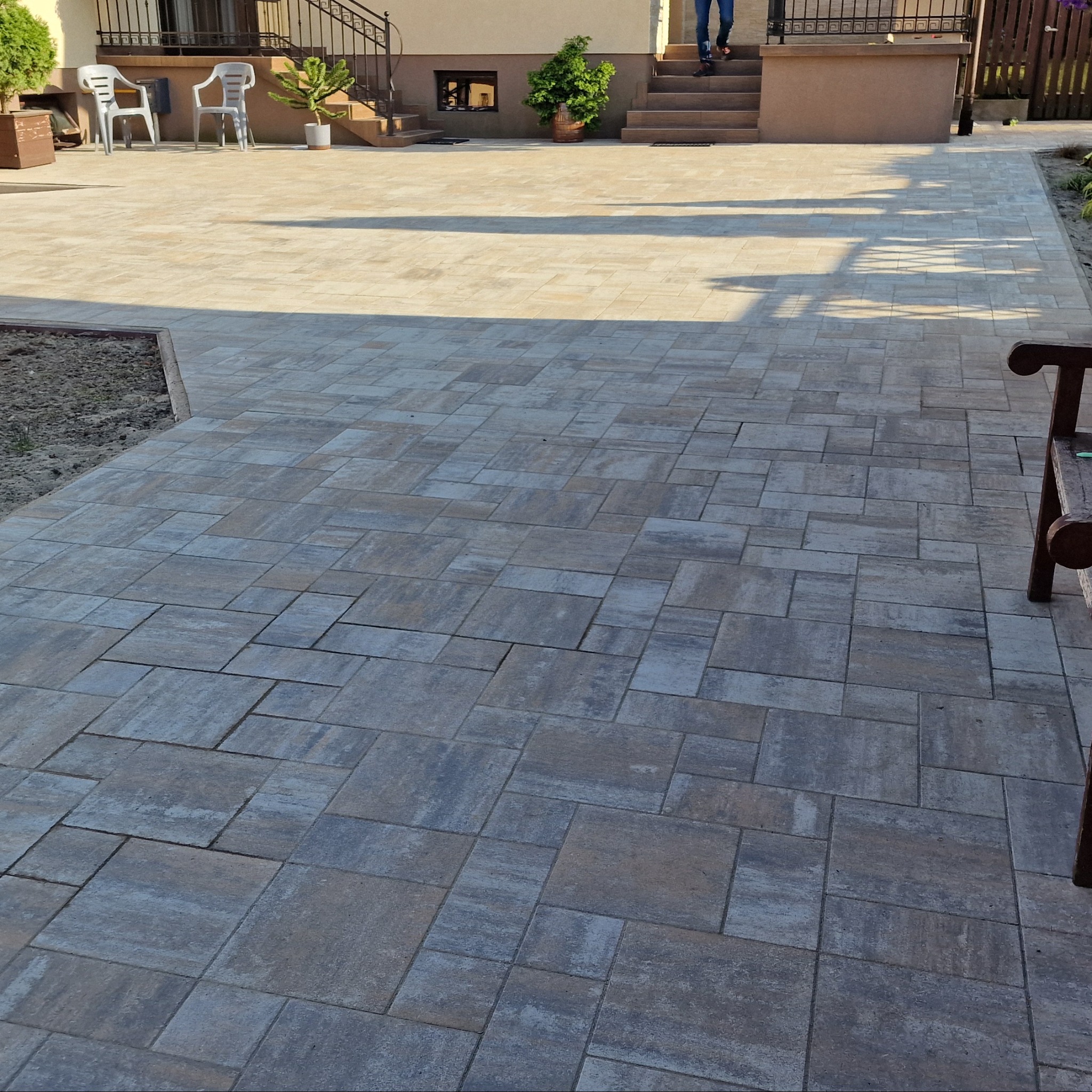 Ułożona kostka brukowa w odcieniach szarości i beżu tworzy wzorzysty chodnik przed domem. Widoczne schody wejściowe i elementy architektury ogrodowej.