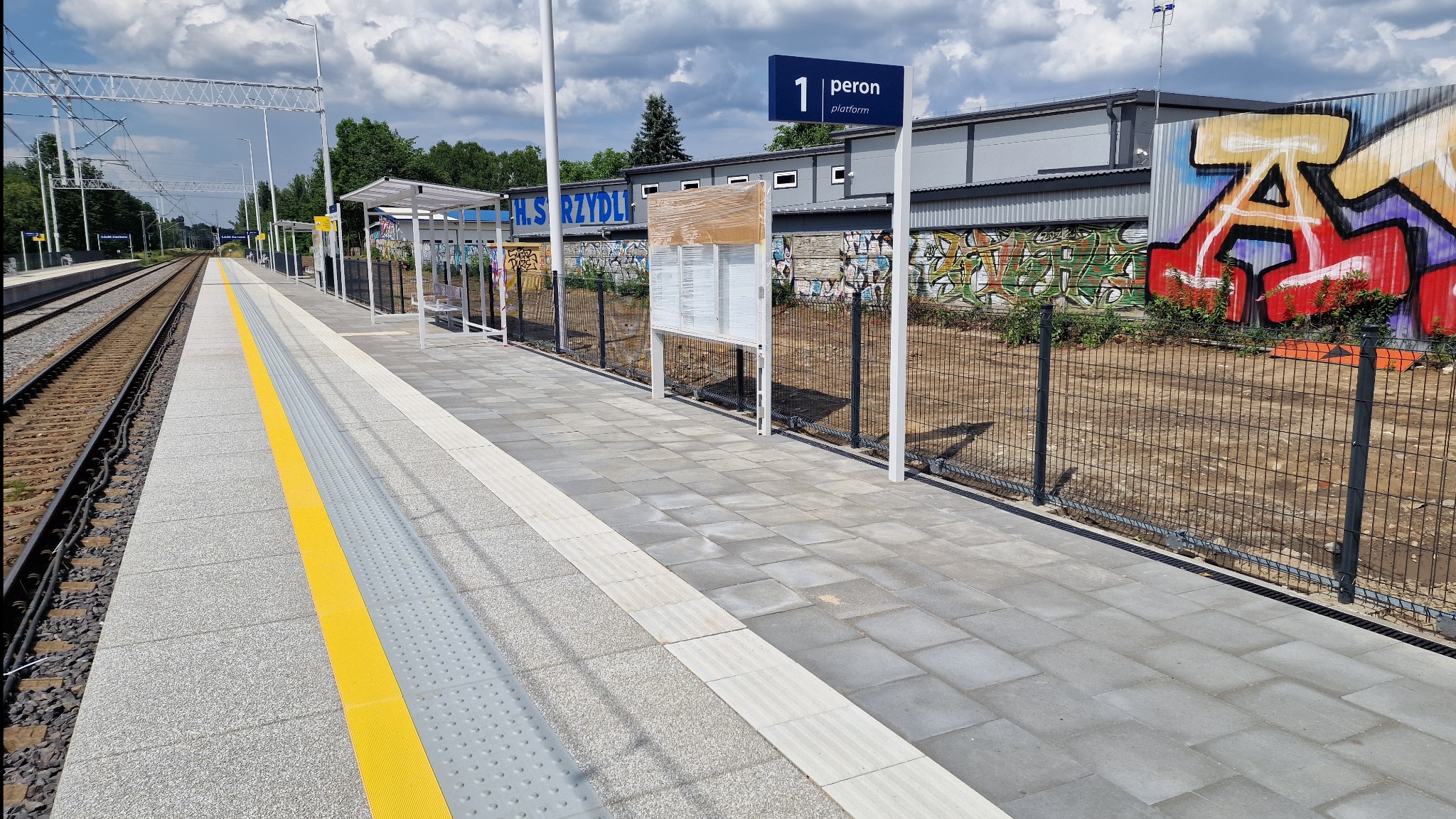 Nowy peron z kostki brukowej z żółtymi pasami ostrzegawczymi, metalowe wiaty i tablice informacyjne. W tle budynek z graffiti i linia kolejowa.