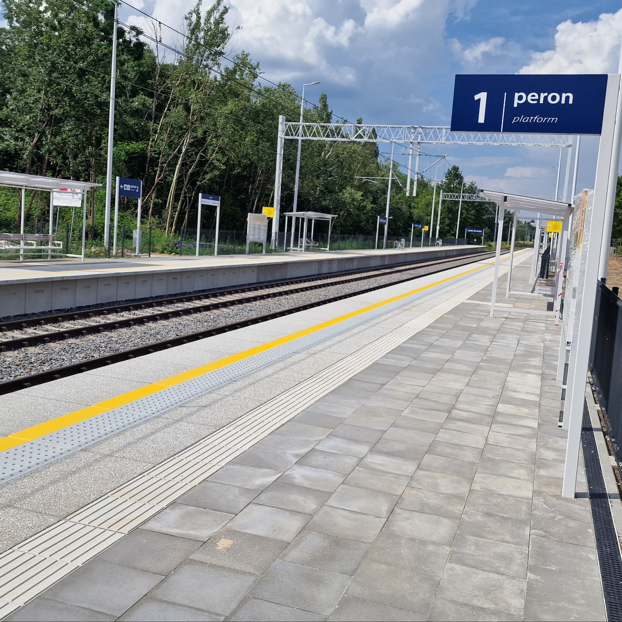 Nowy peron z kostki brukowej, żółte pasy bezpieczeństwa i oznaczenia. Widok na tory i zadaszone wiaty. Słoneczny dzień, błękitne niebo z chmurami.