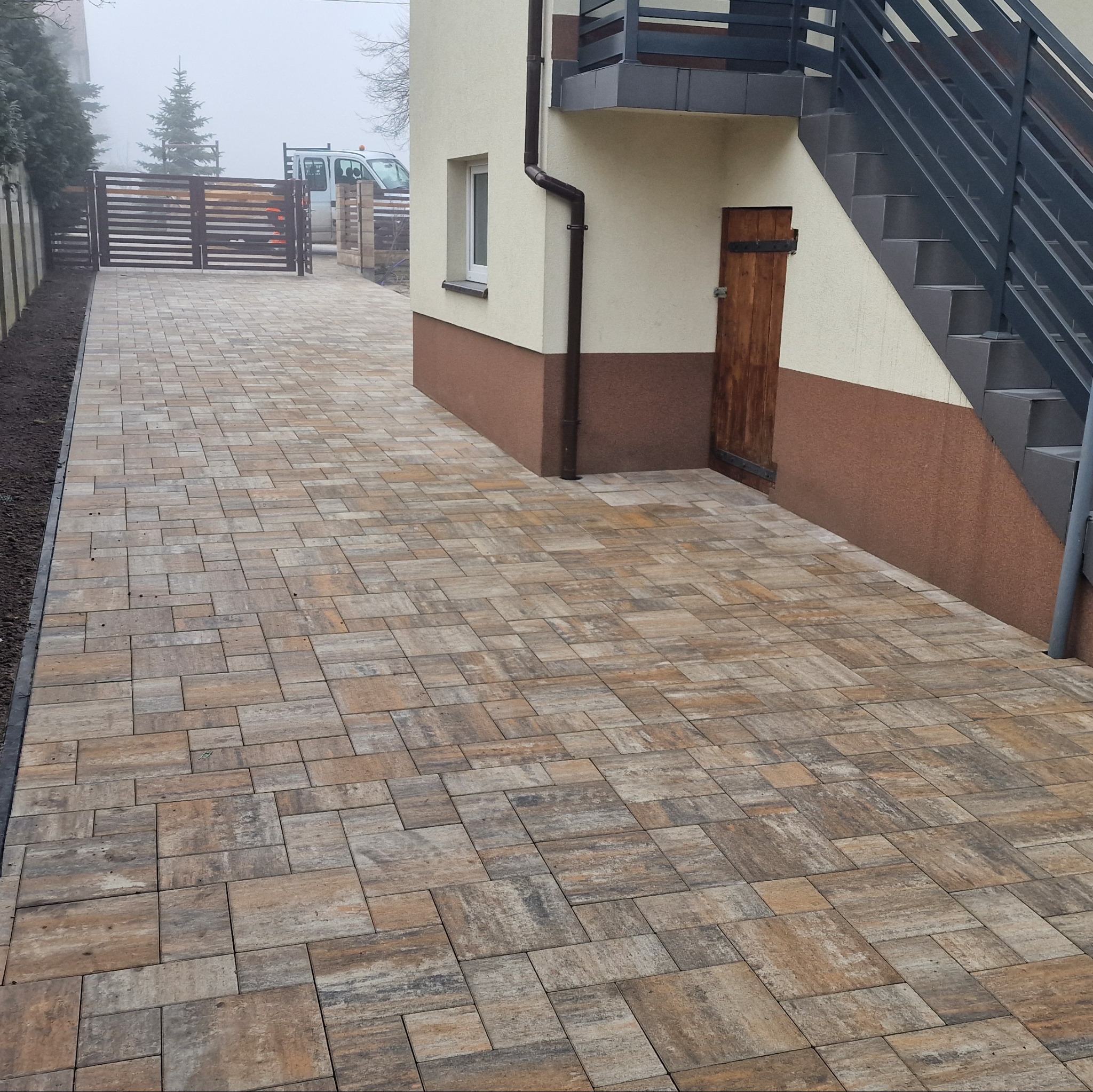 Ułożona kostka brukowa w Skierniewicach, prowadząca do bramy wjazdowej. Widoczny fragment budynku z zewnętrznymi schodami i drewnianymi drzwiami.