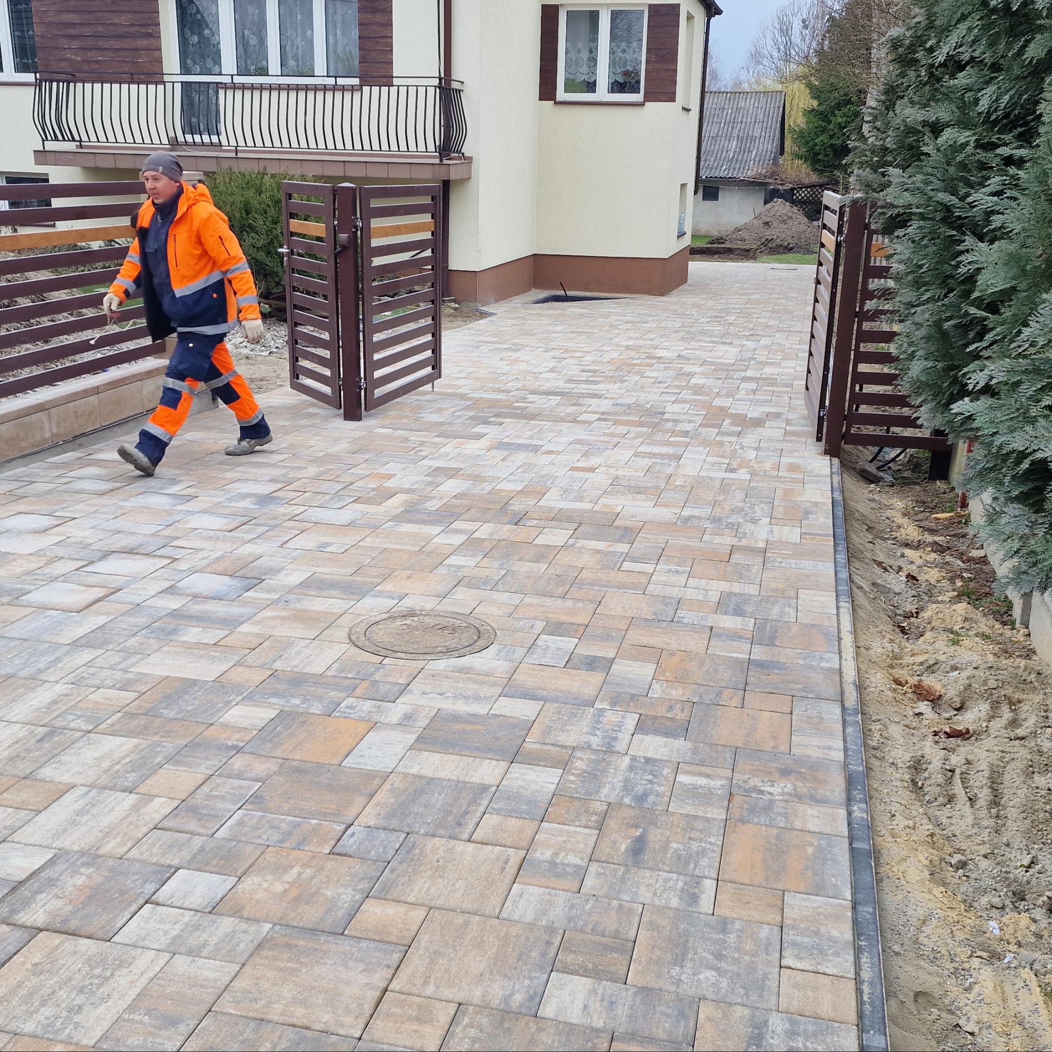 Ułożona kostka brukowa na podjeździe z otwartą bramą; widoczny pracownik w odblaskowym ubraniu, budynek z balkonem i fragment ogrodu z iglakami.
