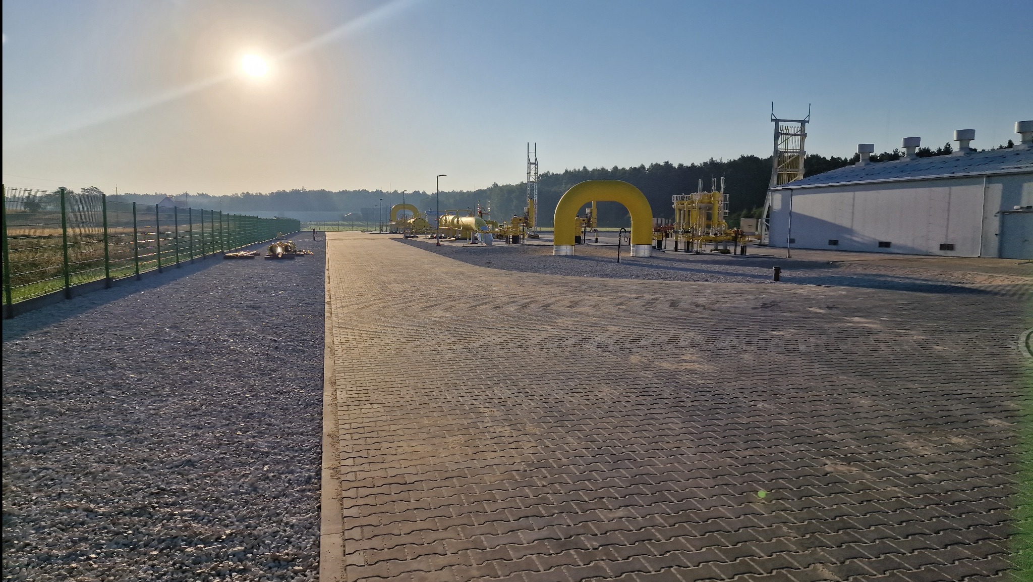 Szeroki plac wyłożony szarą kostką brukową, z żółtymi elementami infrastruktury gazowej w tle, oświetlony wschodzącym słońcem. Długi, zielony płot wzdłuż żwirowej ścieżki po lewej.
