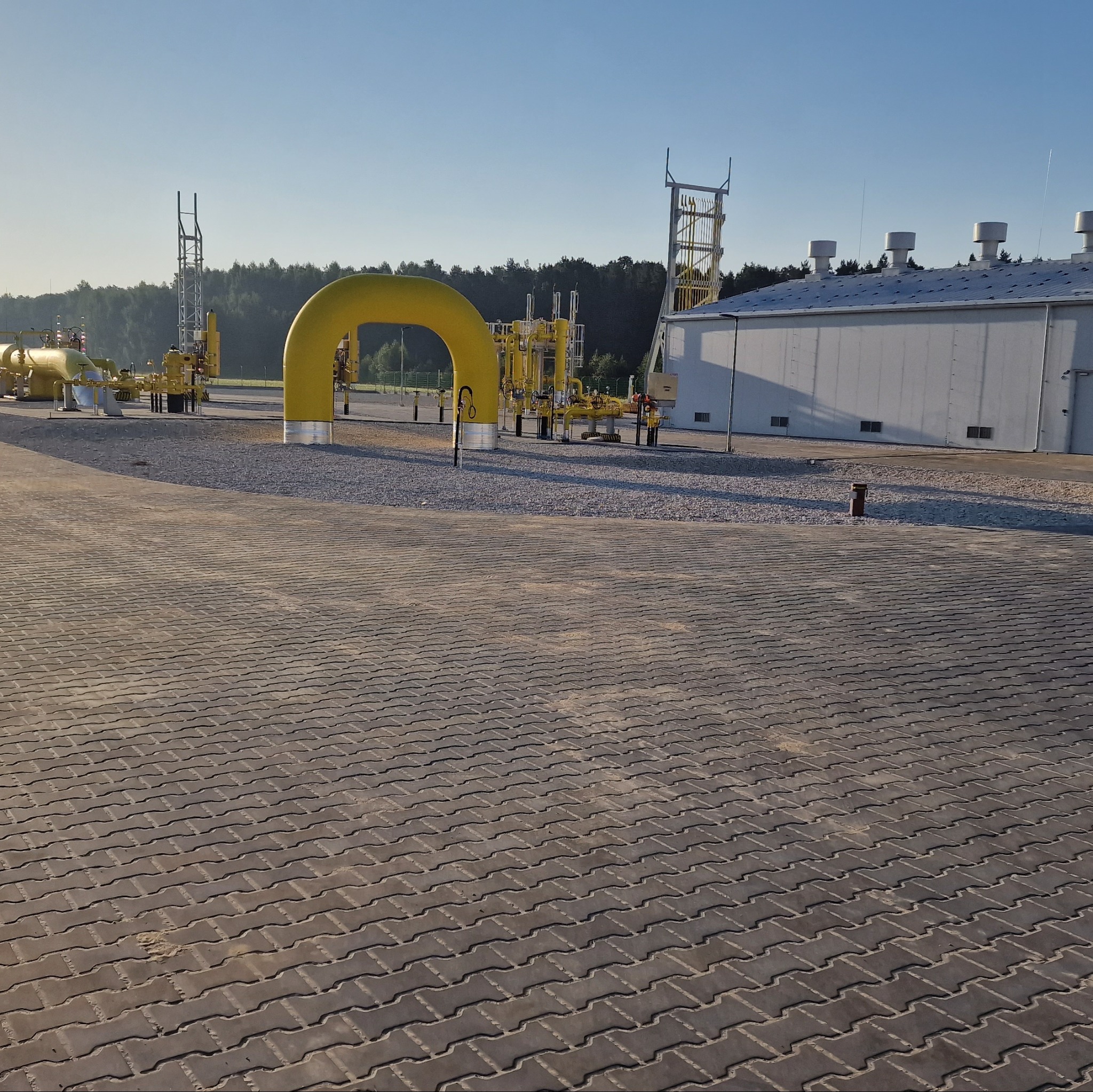 Szeroki kadr na szarą kostkę brukową prowadzącą do żółtej instalacji przemysłowej i budynku w tle, przy słonecznym dniu.