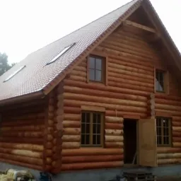 Nowo wybudowany, drewniany dom z bali z czerwoną dachówką, widoczny z zewnątrz w pochmurny dzień, z otwartymi drzwiami i niezagospodarowanym terenem wokół.