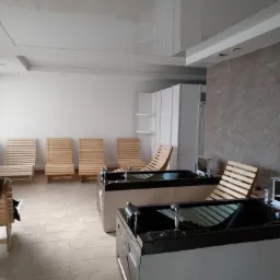 Wnętrze pomieszczenia z dwoma wannami z hydromasażem i drewnianymi leżakami, jasne płytki podłogowe, błyszczący sufit podwieszany, minimalistyczny design.