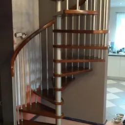 Spiralne schody z drewnianymi stopniami i metalowymi balustradami, widoczne narzędzia na podłodze, wnętrze domu.