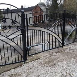 Parys Paweł - Czarna, metalowa brama wjazdowa z automatyką, zdobiona motywem liści, częściowo otwarta, na tle szarej nawierzchni i budynku z czerwonej cegły.