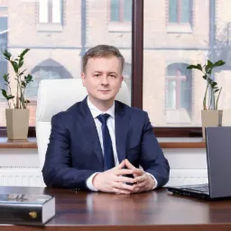 Portret mężczyzny w garniturze siedzącego za biurkiem z laptopem i książkami, na tle okna z widokiem na budynek z cegły.