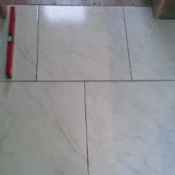 Układanie jasnych, marmurkowych płytek podłogowych z użyciem czerwonej poziomicy i krzyżyków dystansowych, widoczne ślady zaprawy.