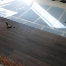 Układanie ciemnych paneli podłogowych, widoczna podkładka z taśmą, młotek i kątownik w słonecznym wnętrzu.