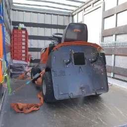 Traktorek do koszenia trawy marki Stihl zabezpieczony pasami wewnątrz przestrzeni ładunkowej busa, obok widoczne czerwone skrzynki i szare koce.