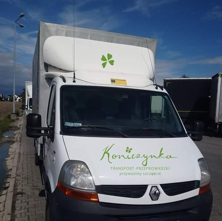 Biały samochód dostawczy z logo firmy transportowej w kształcie zielonej koniczyny, zaparkowany na parkingu z widocznymi innymi pojazdami w tle.