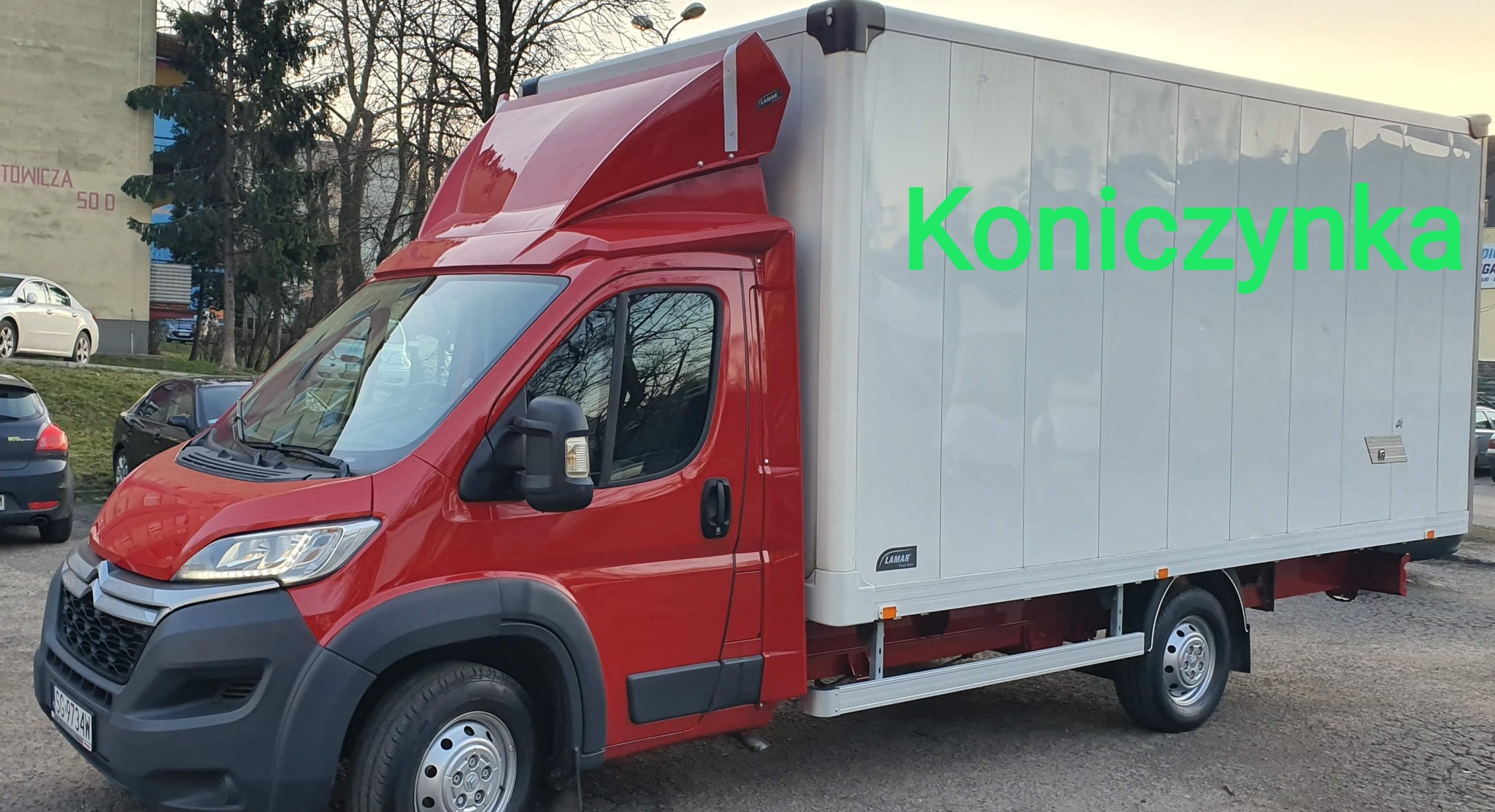 Czerwony bus dostawczy marki Citroen z białą zabudową kontenerową, zaparkowany na tle budynku z napisem 'Towicza 500'.