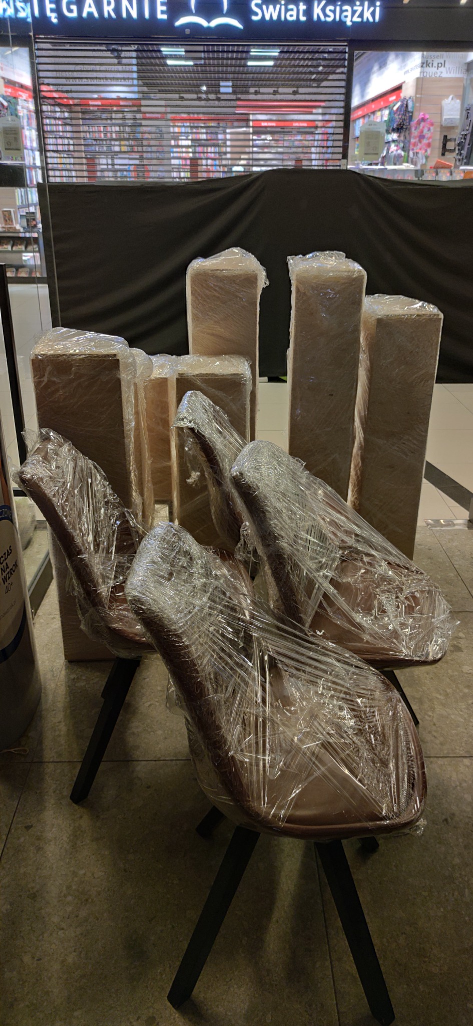Krzesła i kartony zabezpieczone folią stretch, przygotowane do transportu wewnątrz galerii handlowej z zamkniętymi sklepami.