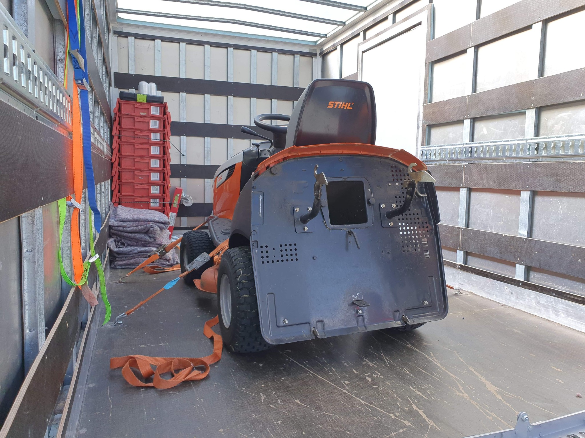 Traktorek do koszenia trawy marki Stihl zabezpieczony pasami wewnątrz przestrzeni ładunkowej busa, obok widoczne czerwone skrzynki i szare koce.