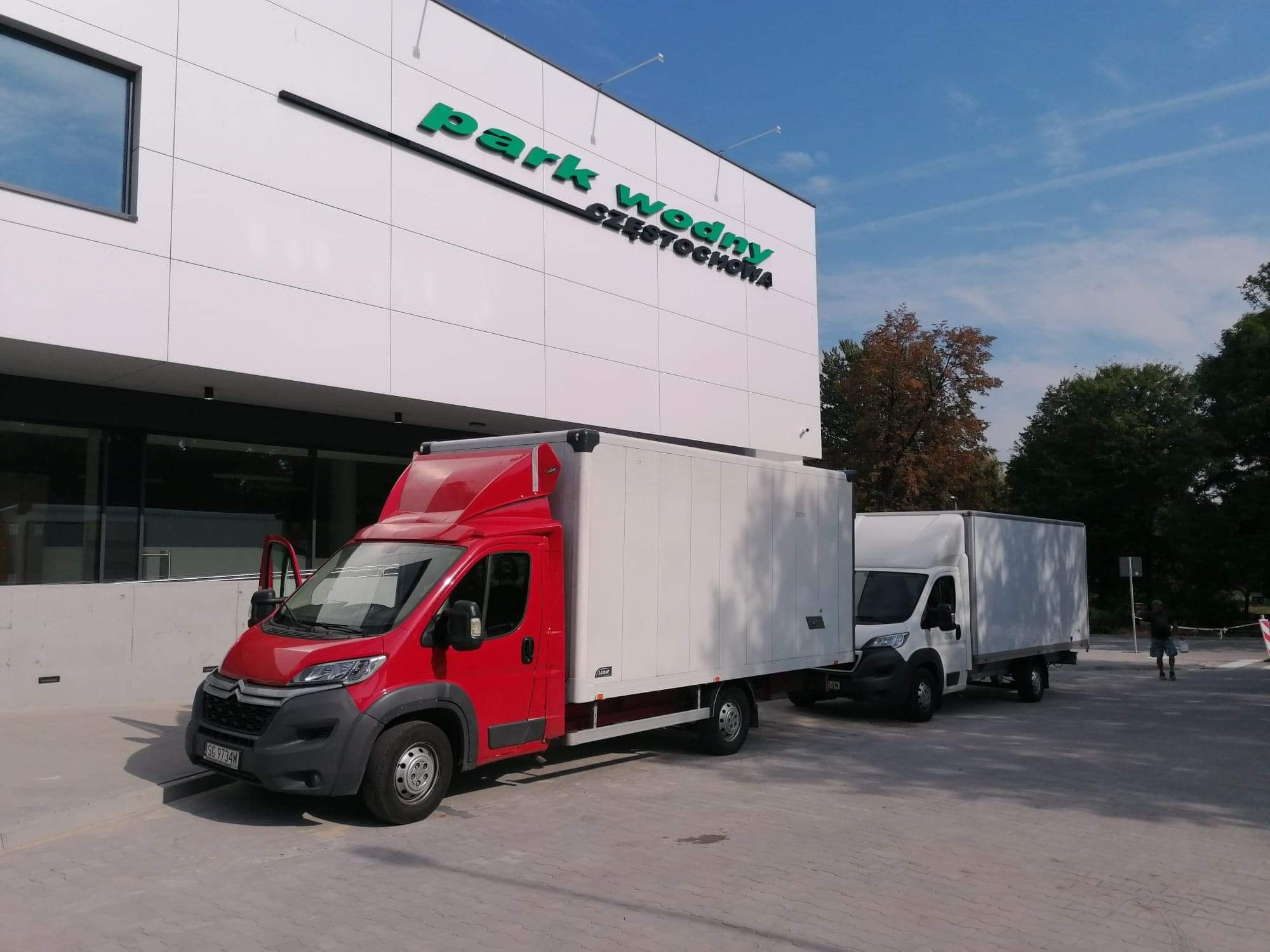 Dwa busy transportowe, czerwony i biały, zaparkowane przed budynkiem Parku Wodnego Częstochowa w słoneczny dzień. Czerwony bus ma otwarte drzwi kierowcy.