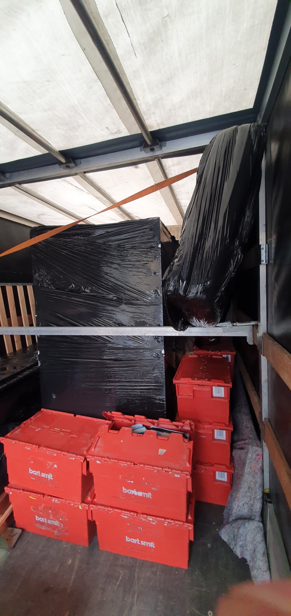 Wnętrze przestrzeni ładunkowej busa z zabezpieczonymi meblami owiniętymi folią i stosem czerwonych plastikowych skrzyń z napisem 'bart smit'.