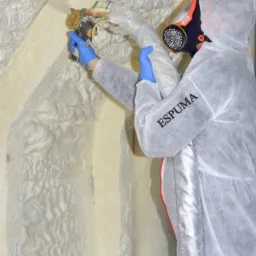 Pracownik w kombinezonie ochronnym i masce natryskuje izolację na ścianę, widoczna struktura natryskiwanej piany.