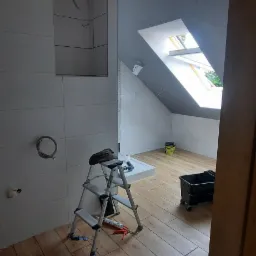 Łazienka na poddaszu w trakcie remontu: białe płytki ścienne, drewnopodobna podłoga, okno dachowe, drabina malarska z narzędziami, widoczne przygotowania do montażu armatury.