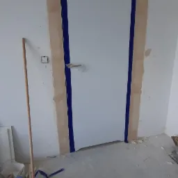Białe drzwi wewnętrzne z niebieską taśmą malarską, osadzone w ościeżnicy z widocznymi śladami gładzi szpachlowej, na podłodze leży szczotka i drabina