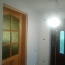 Wnętrze po remoncie z drewnianą futryną i drzwiami z szybami, widoczne kable wystające z sufitu, świeżo pomalowane ściany i szara podłoga.