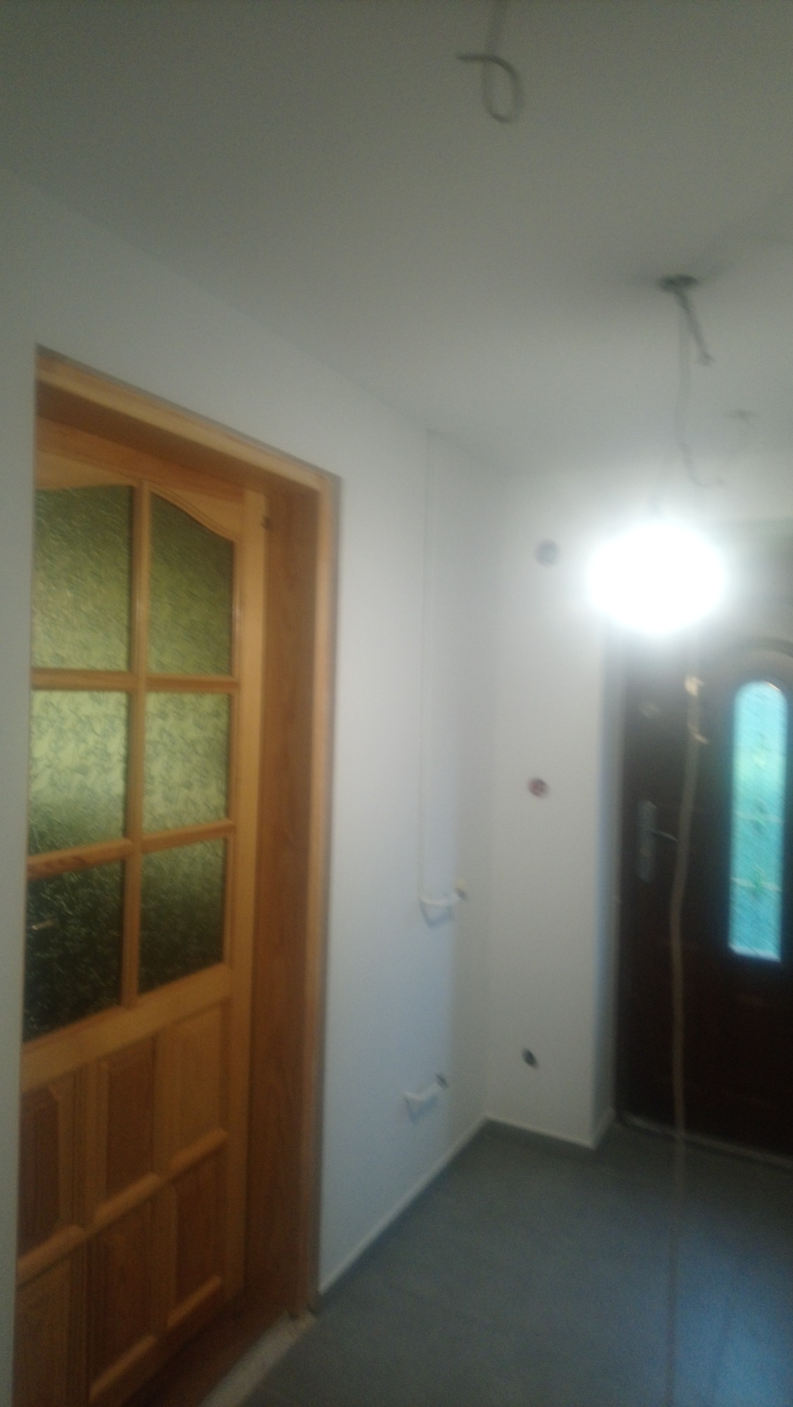 Wnętrze po remoncie z drewnianą futryną i drzwiami z szybami, widoczne kable wystające z sufitu, świeżo pomalowane ściany i szara podłoga.