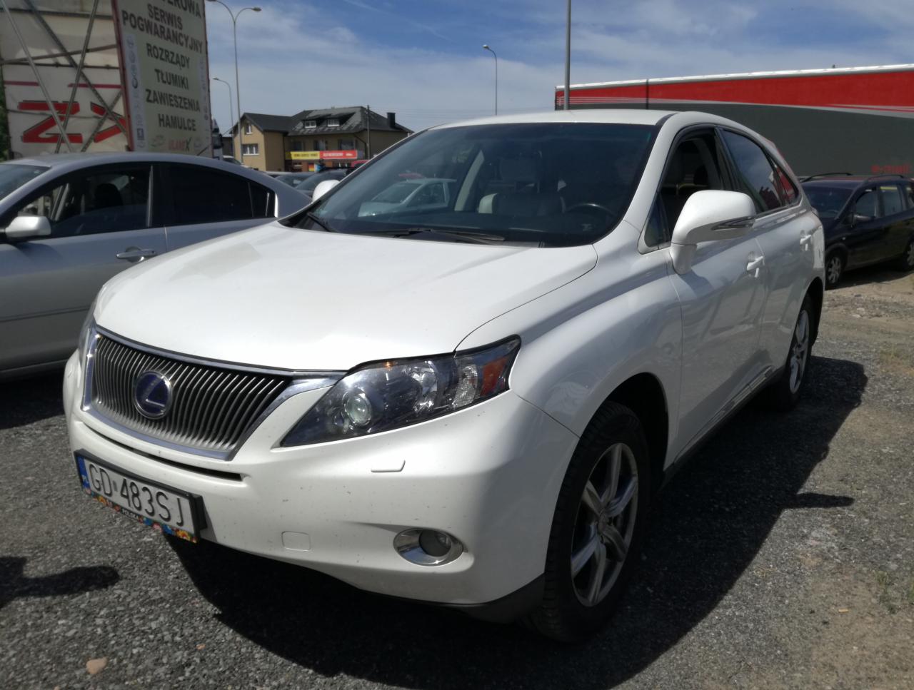 Biały samochód Lexus SUV na parkingu, widoczny z przodu, tablica rejestracyjna GD 483SJ, w tle budynki i inne pojazdy.